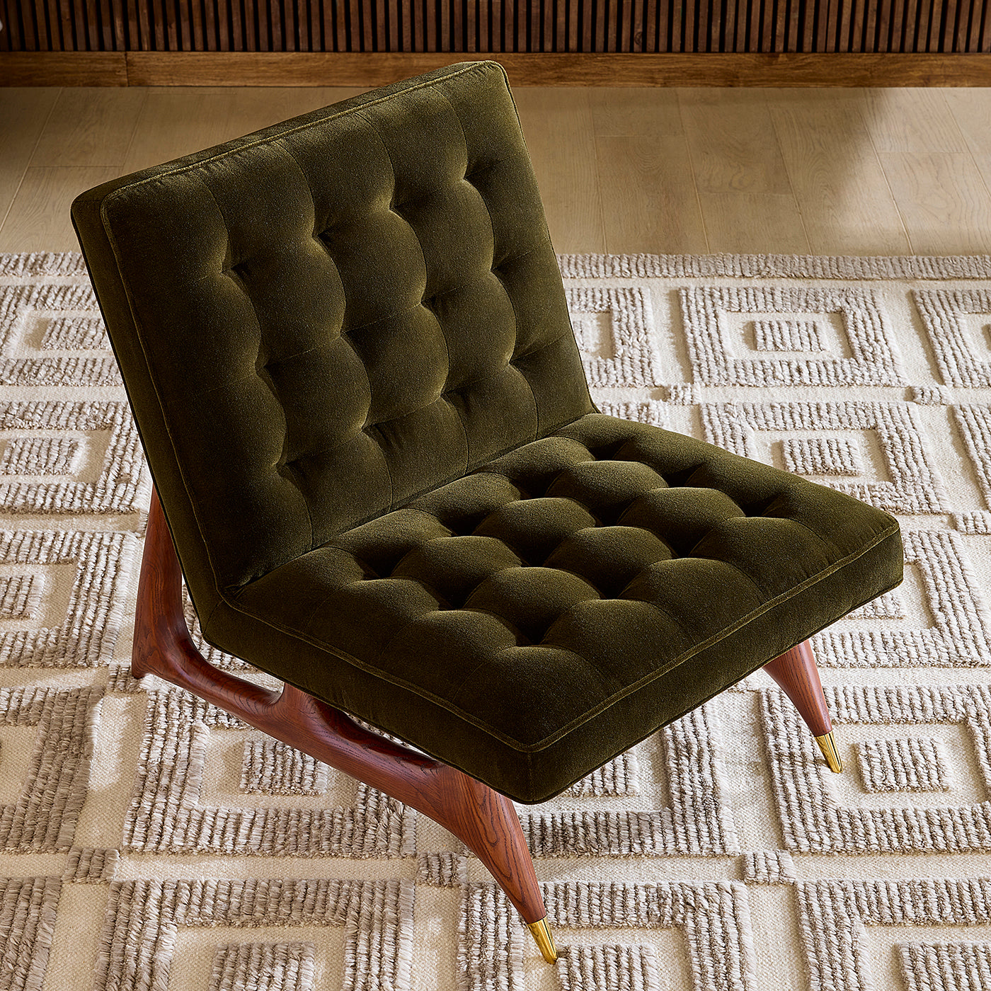An olive green tufted velvet accent chair with a wooden frame and gold-toned metal legs, positioned on a textured beige rug in a modern interior setting.