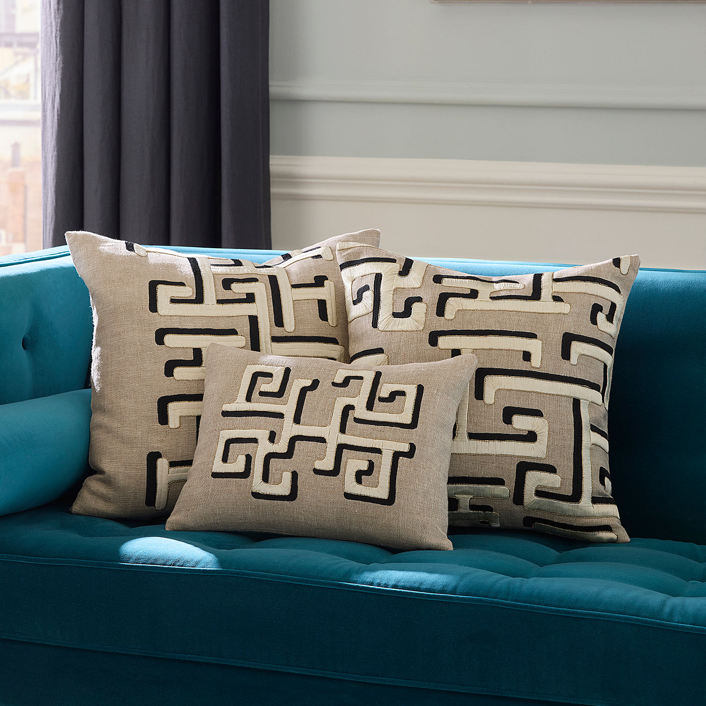 Three beige throw pillows with a black Greek key pattern arranged on a teal tufted sofa in a living room setting.