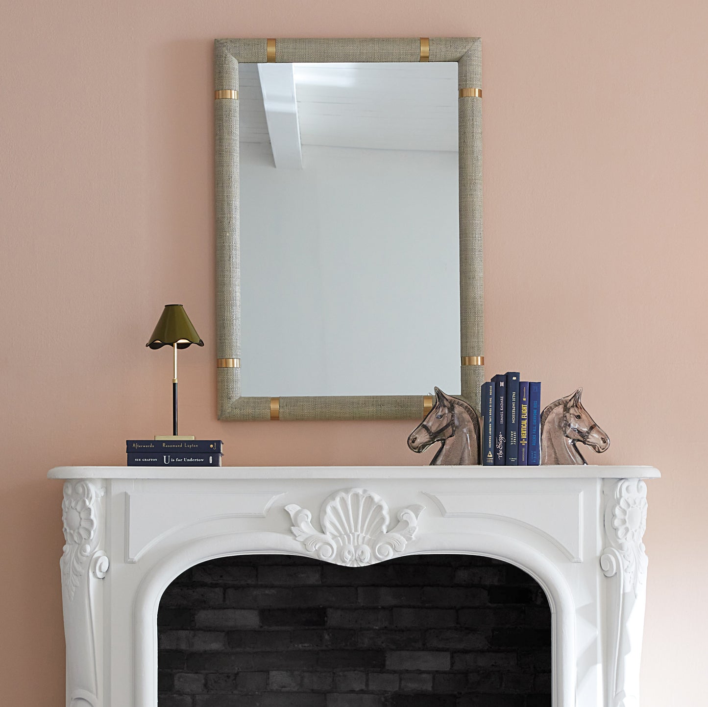A rectangular wall mirror with a textured beige linen frame and gold corner accents, mounted on a dusty pink wall above a white fireplace mantel styled with books and horse figurines.