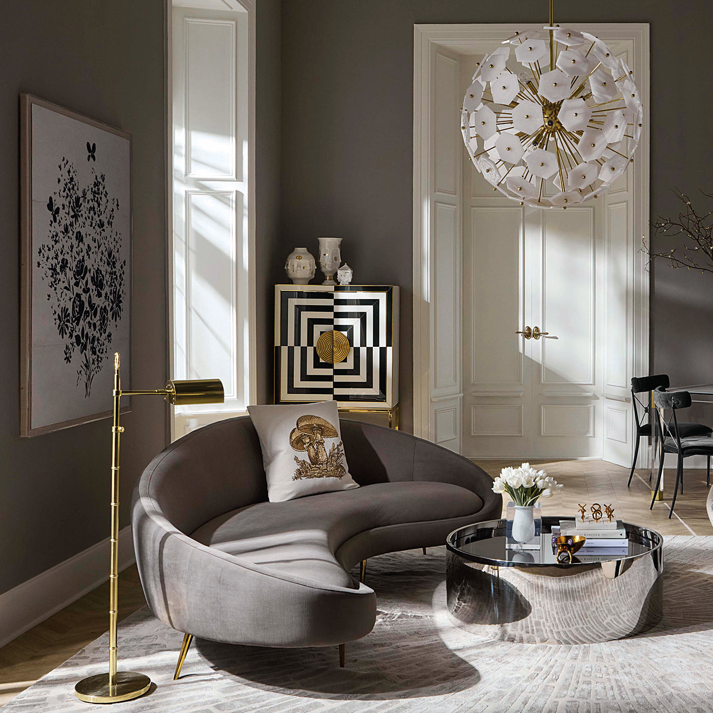 A modern curved gray velvet sofa in a styled living room with a gold floor lamp, a round mirrored coffee table, a white floral chandelier, and dark gray walls.