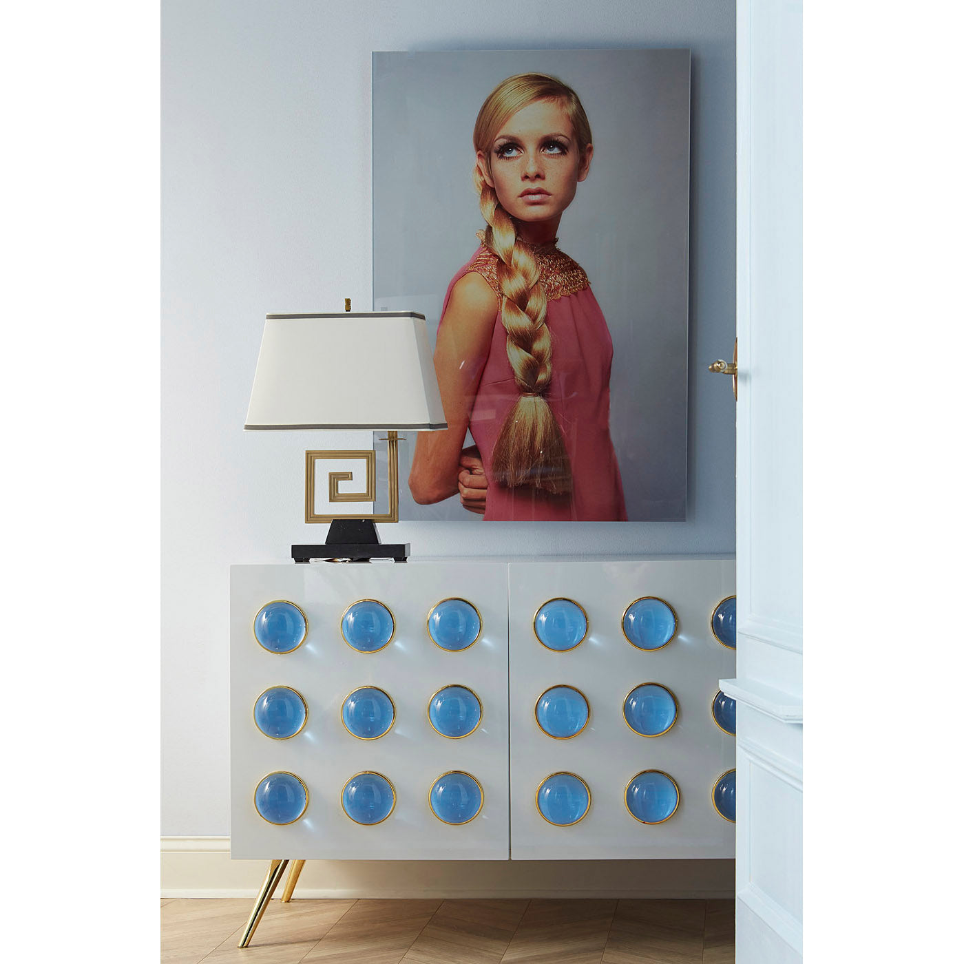 Decorative cabinet with blue circular accents and a lamp, with a portrait of a woman on the wall.