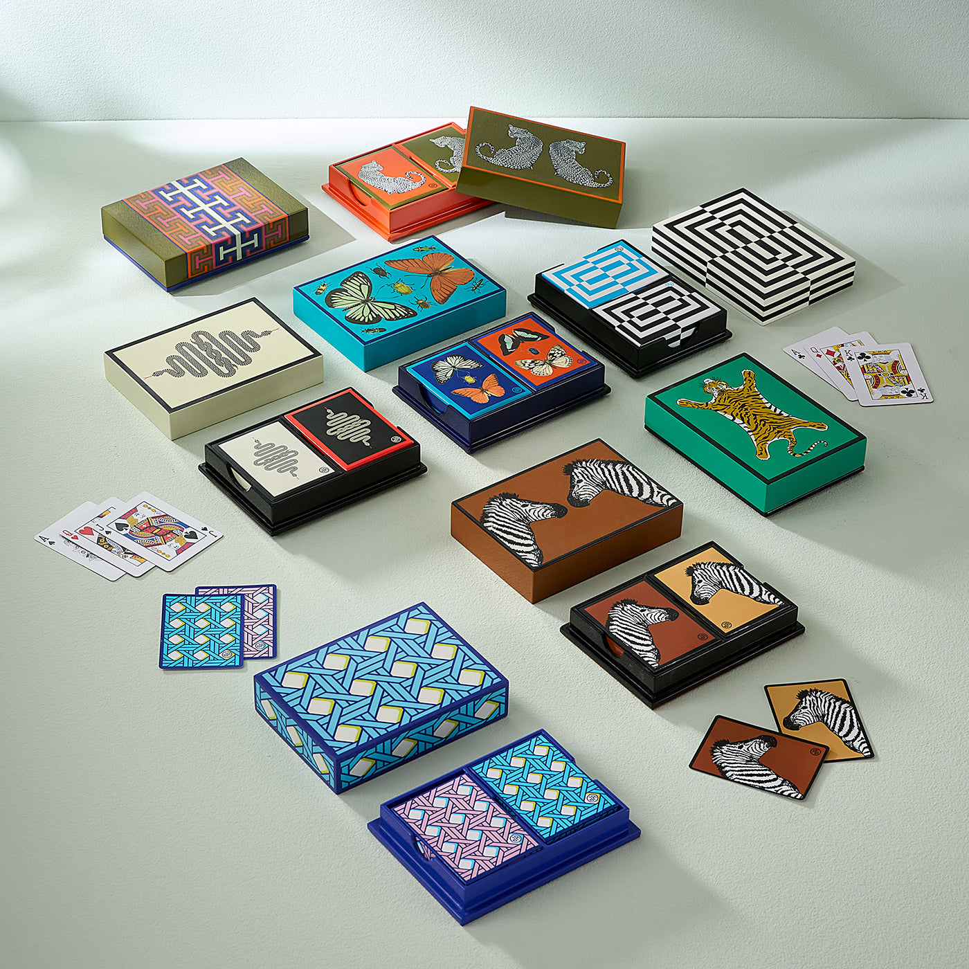 An assortment of colorful playing cards and matching decorative boxes arranged on a light gray surface. The sets feature various patterns including zebra prints, stripes, geometric designs, butterflies, and abstract shapes in blue, green, orange, black, white, and brown.
