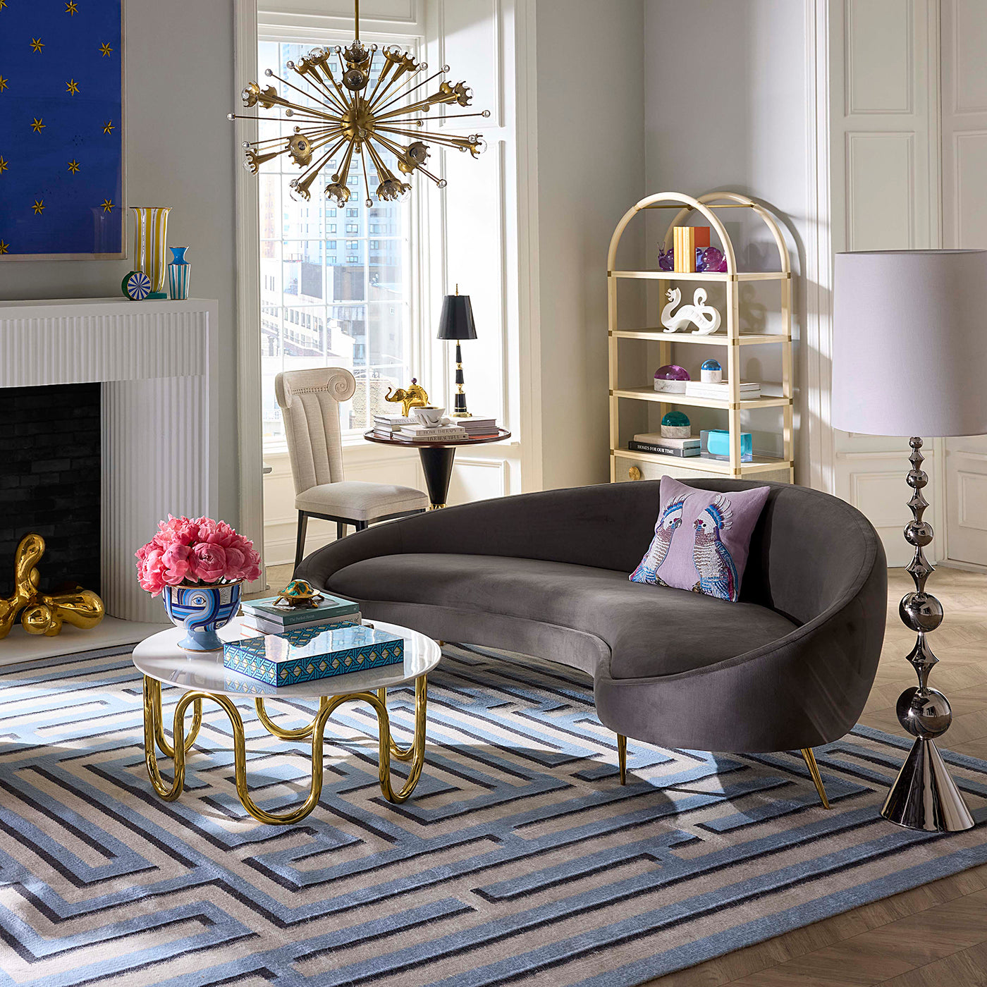 A modern curved velvet sofa in dark taupe-gray sits in a styled living room with a blue patterned rug, gold coffee table, pink flowers, shelving, and a window view.