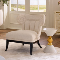 An upholstered accent chair in a light beige fabric with black legs, positioned next to a white and gold pedestal side table in a bright, modern room with wood floors and large windows.
