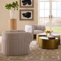 A styled living room featuring a light beige upholstered armchair in the foreground, a matching sofa behind it, two round metallic gold coffee tables, framed wall art, a plant on a pedestal, and tall windows with a view of greenery outside.