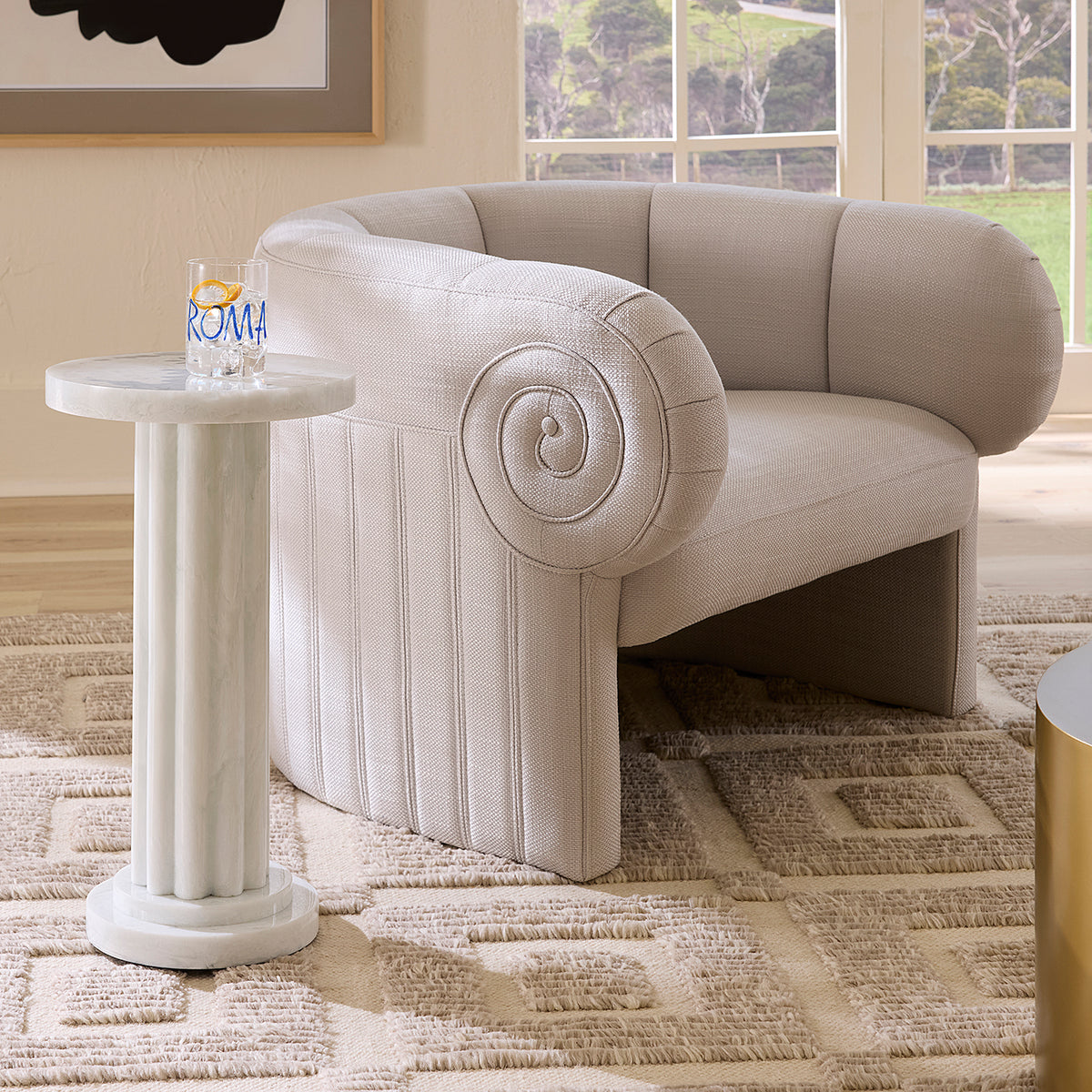 A light beige upholstered accent chair with a flared base and spiral arm detail, shown in a styled room next to a white column side table.