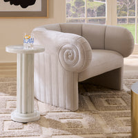 A light beige upholstered accent chair with a flared base and spiral arm detail, shown in a styled room next to a white column side table.
