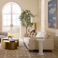 A styled living room featuring a cream bouclé sofa with patterned throw pillows, a white column side table, round metallic gold coffee tables, a patterned rug, and a large arched window in the background.