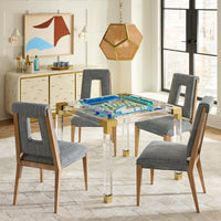 A modern dining set featuring a clear acrylic table with gold metal accents and four gray upholstered chairs with light wood legs, styled in a bright, contemporary dining room.
