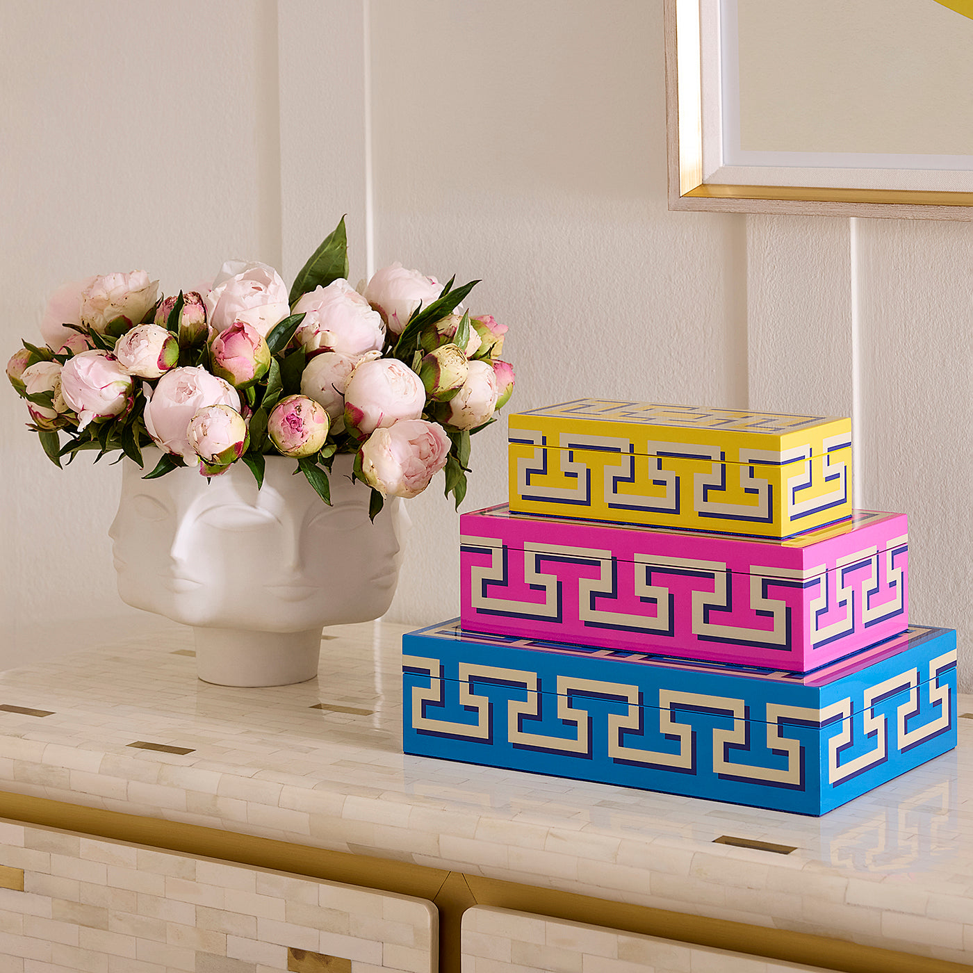 Three stacked decorative storage boxes with a Greek key pattern in blue, pink, and yellow, styled on a console table next to a white ceramic vase of pink peonies.