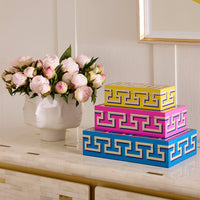 Three stacked decorative storage boxes with a Greek key pattern in blue, pink, and yellow, styled on a console table next to a white ceramic vase of pink peonies.