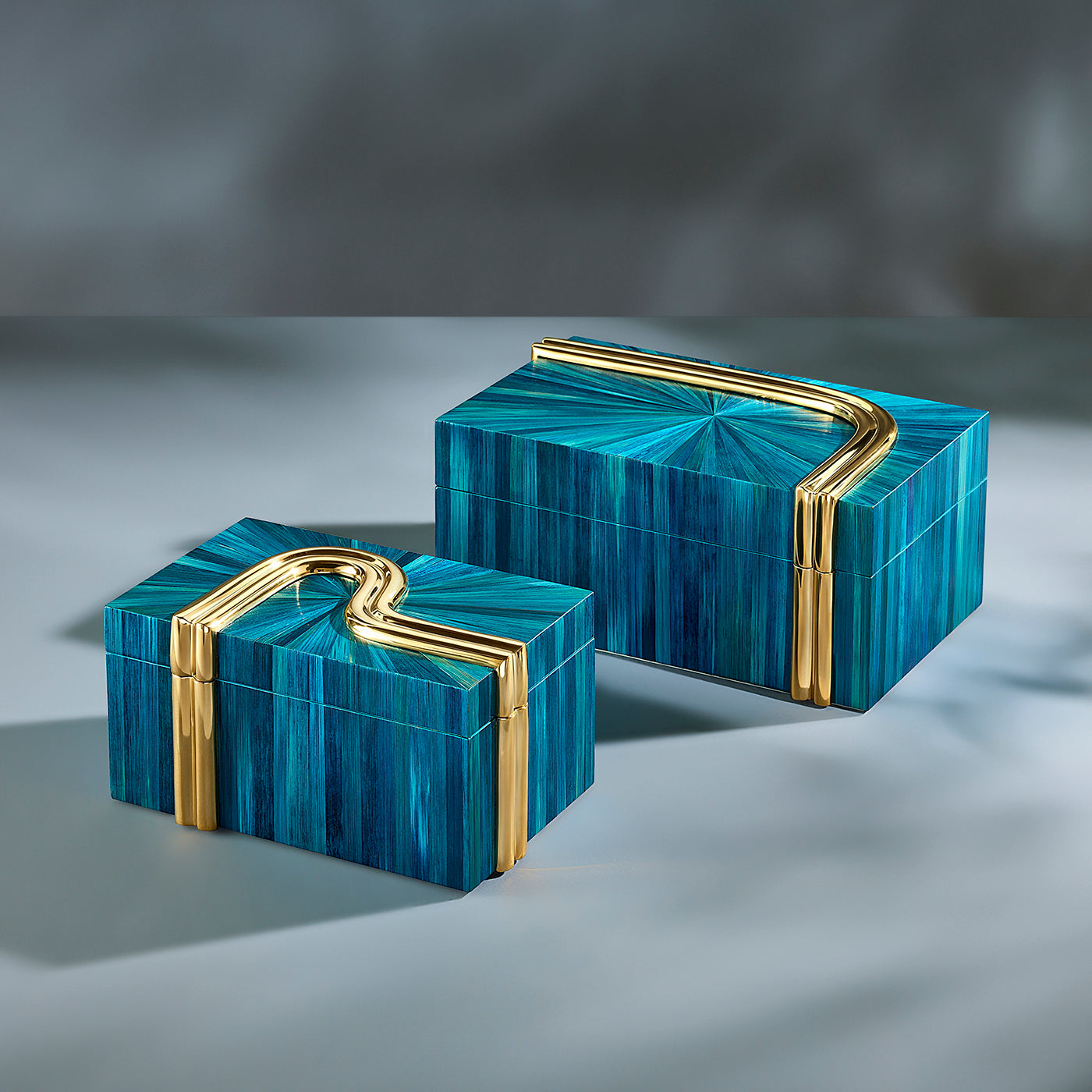 Two rectangular lidded boxes with geometric blue striped inlay and polished gold metal handles, arranged on a gray studio surface.