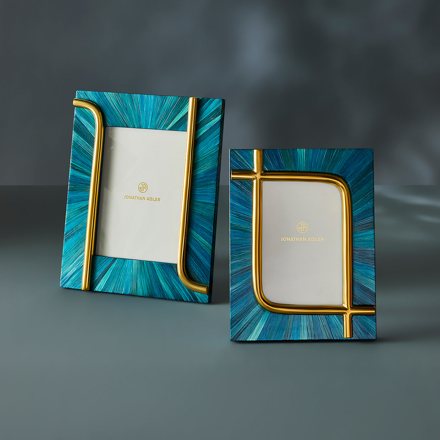 Two Jonathan Adler picture frames with a blue and teal sunburst pattern and gold metallic accents, displayed on a dark gray surface.