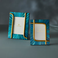 Two Jonathan Adler picture frames with a blue and teal sunburst pattern and gold metallic accents, displayed on a dark gray surface.