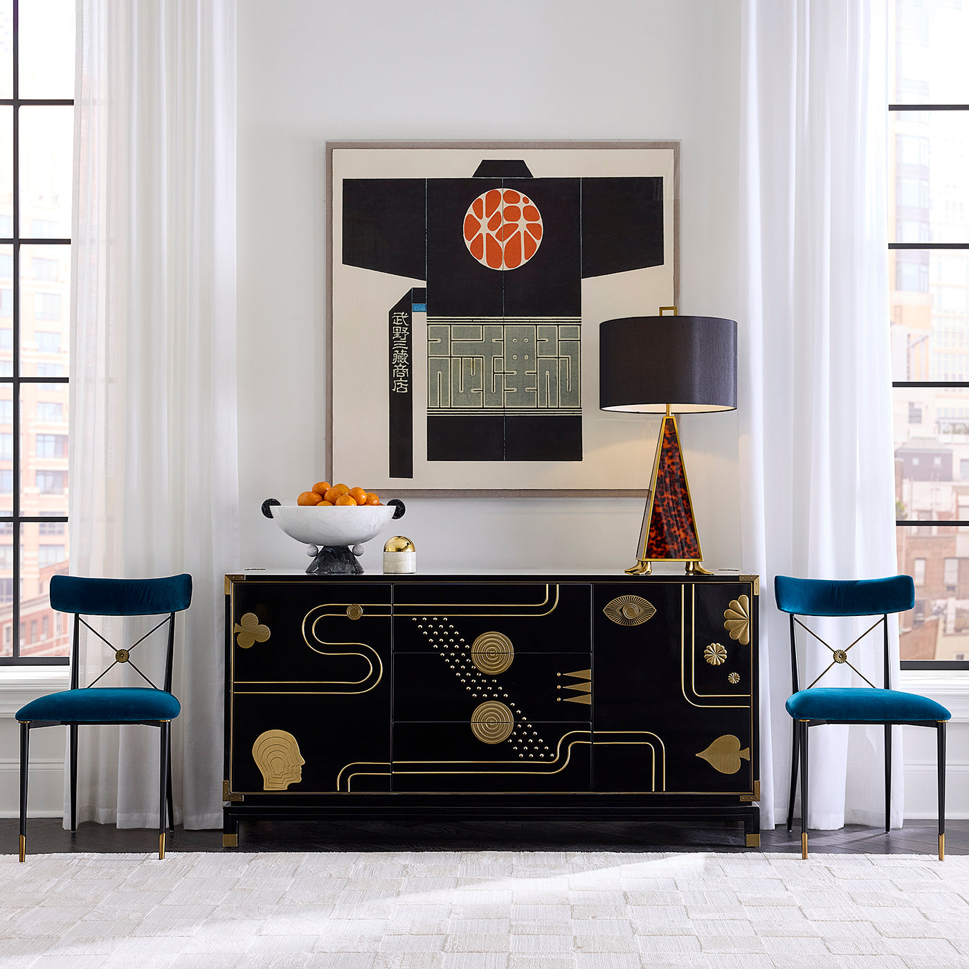 A black Art Deco style sideboard cabinet with gold geometric patterns, styled in a bright modern room with two teal velvet chairs, a table lamp, and a framed abstract print on the wall.