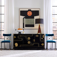 A black Art Deco style sideboard cabinet with gold geometric patterns, styled in a bright modern room with two teal velvet chairs, a table lamp, and a framed abstract print on the wall.