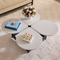 A modern four-segment round coffee table with a speckled light gray stone top and black metal legs, styled in a living room with books, a flower arrangement, and a tic-tac-toe board on top.