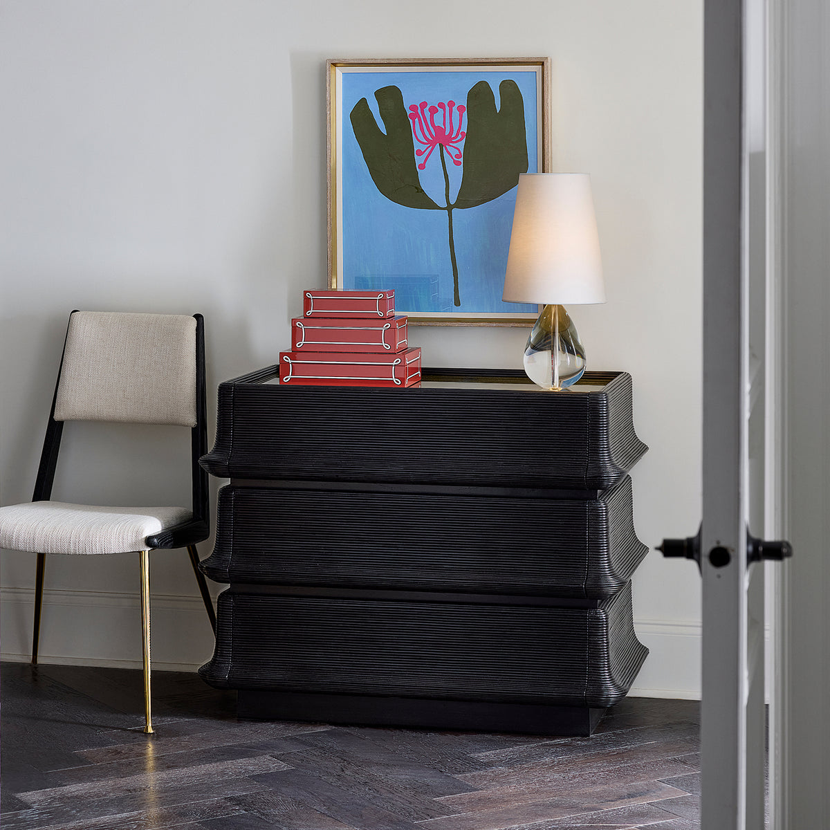 A black textured three-tier chest of drawers styled in a modern room setting, topped with a stack of red books and a glass table lamp with a white shade. A beige upholstered chair sits to the left, and a framed floral art print hangs on the light gray wall above.