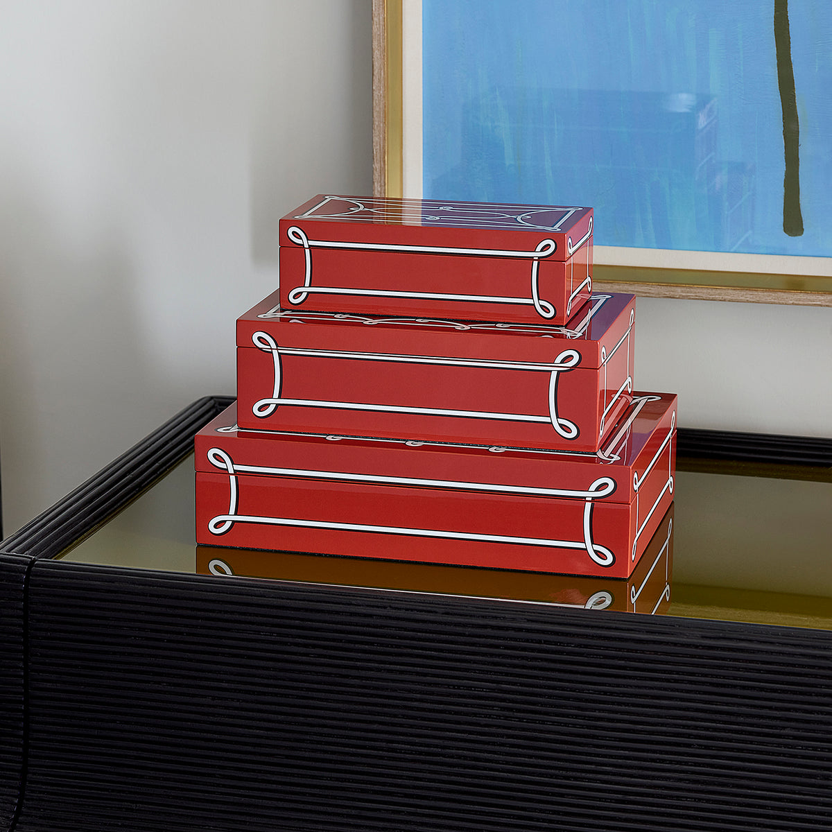 Three stacked red decorative boxes with clear lids and white rope-style trim, displayed on a black ribbed table in an indoor setting.