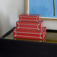 Three stacked red decorative boxes with clear lids and white rope-style trim, displayed on a black ribbed table in an indoor setting.