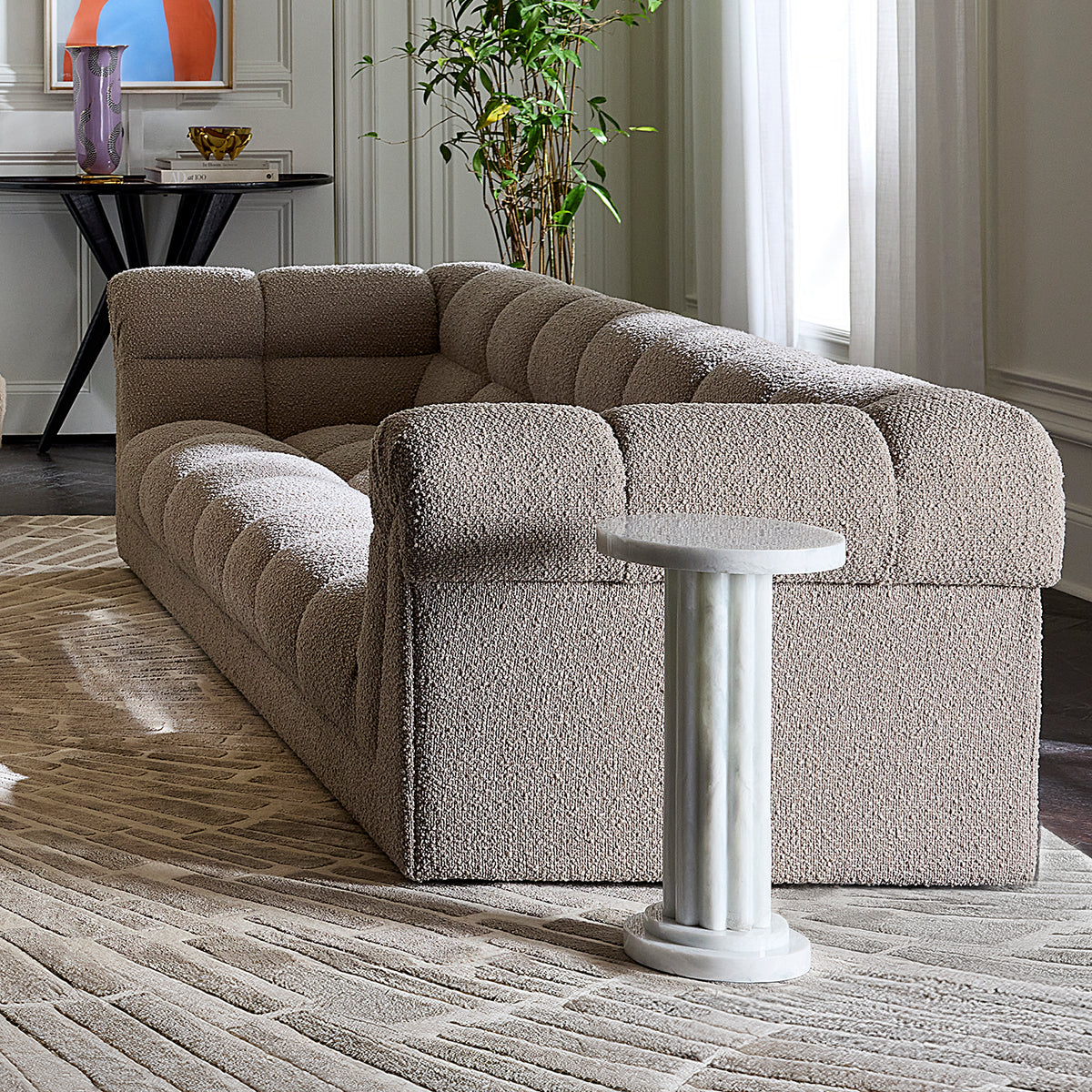 A modern tufted bouclé sofa in a warm taupe-beige color, styled in a bright living room with a white marble side table, textured rug, and potted plant.