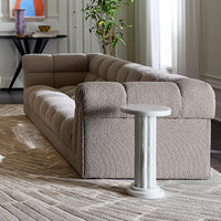 A modern tufted bouclé sofa in a warm taupe-beige color, styled in a bright living room with a white marble side table, textured rug, and potted plant.