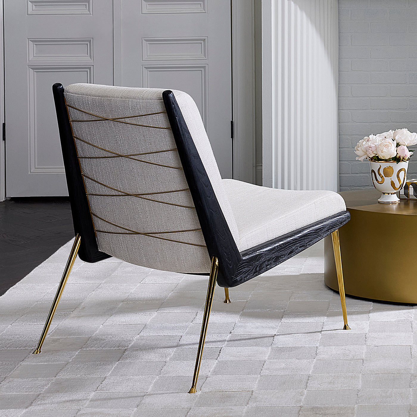 A modern accent chair with a light beige textured fabric seat and back, accented by gold geometric lines, a black side frame, and slim gold metal legs, shown in a styled interior setting.