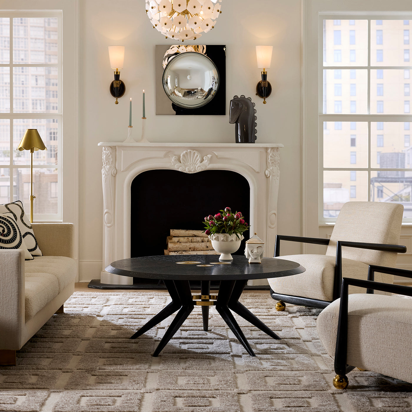 A styled interior living room scene featuring a white carved fireplace mantel, a round black coffee table with black star legs, two cream upholstered armchairs with black frames, a patterned rug, and large windows.