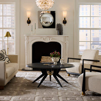 A styled interior living room scene featuring a white carved fireplace mantel, a round black coffee table with black star legs, two cream upholstered armchairs with black frames, a patterned rug, and large windows.