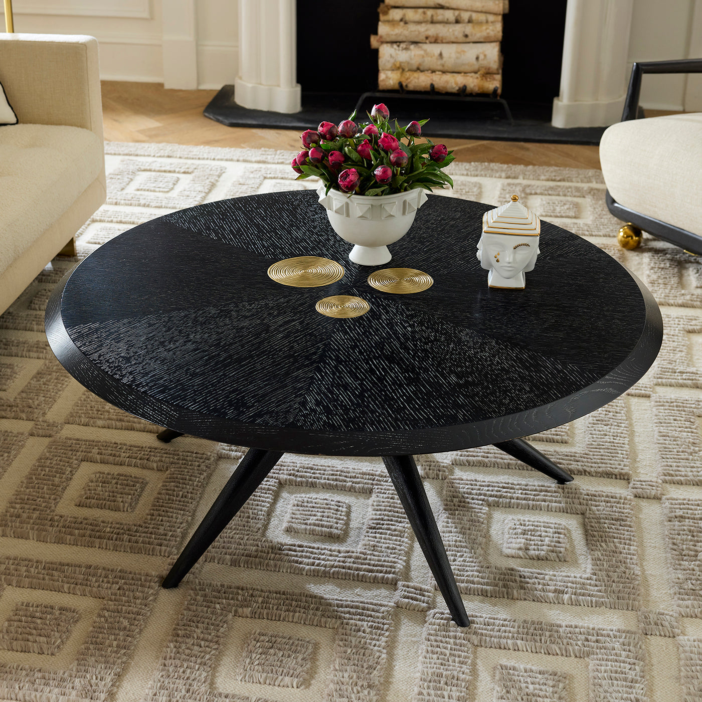 A round black coffee table with a textured wood-grain finish and three angled legs, styled in a living room with a patterned rug, a white floral urn, and decorative accents.