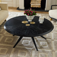 A round black coffee table with a textured wood-grain finish and three angled legs, styled in a living room with a patterned rug, a white floral urn, and decorative accents.