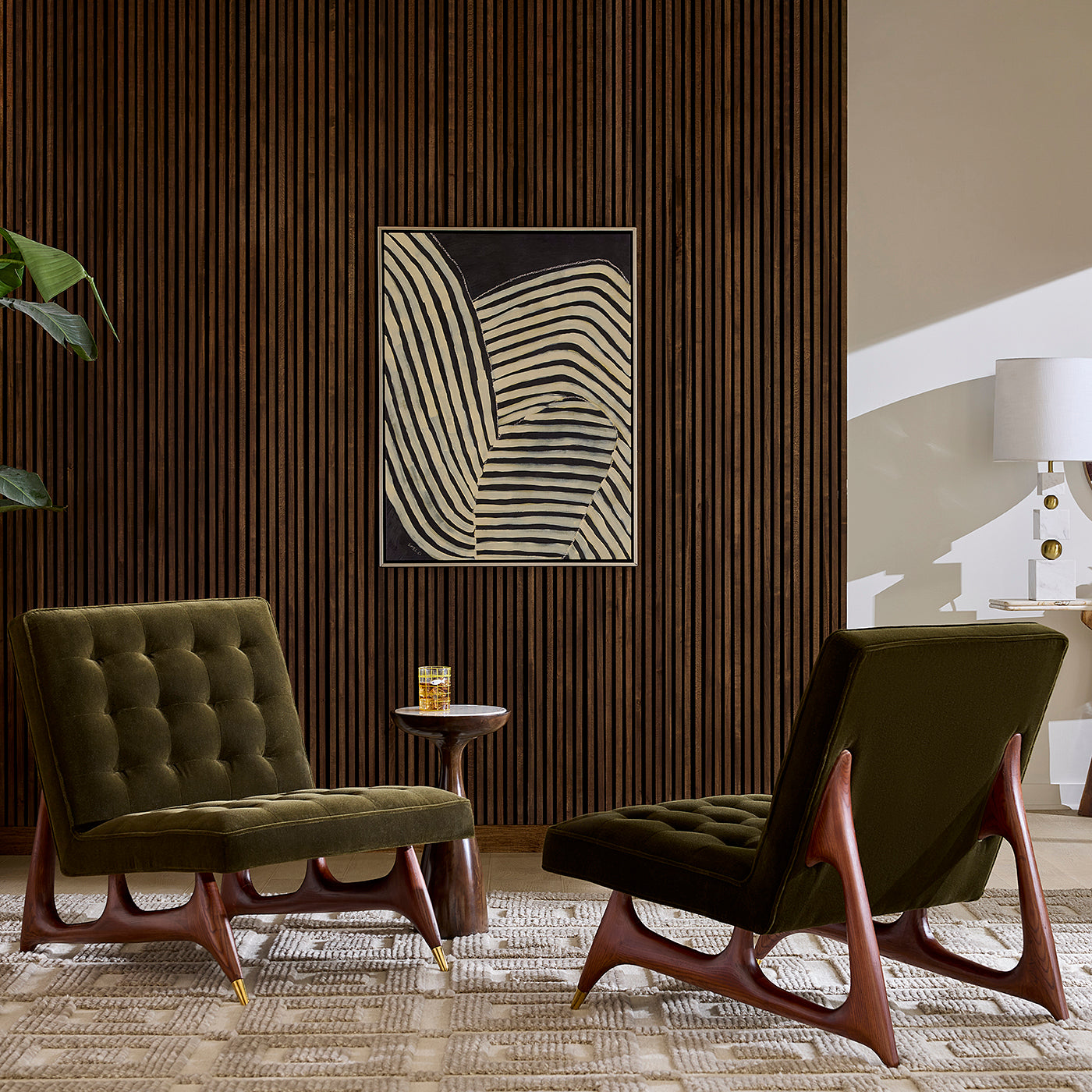 A styled interior scene with two mid-century modern armchairs in dark olive green velvet, set in front of a dark wood slatted wall with a framed abstract art print. A small side table with a drink sits between them, and a lamp is visible on the right.