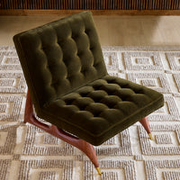 An olive green tufted velvet accent chair with a wooden frame and gold-toned metal legs, positioned on a textured beige rug in a modern interior setting.