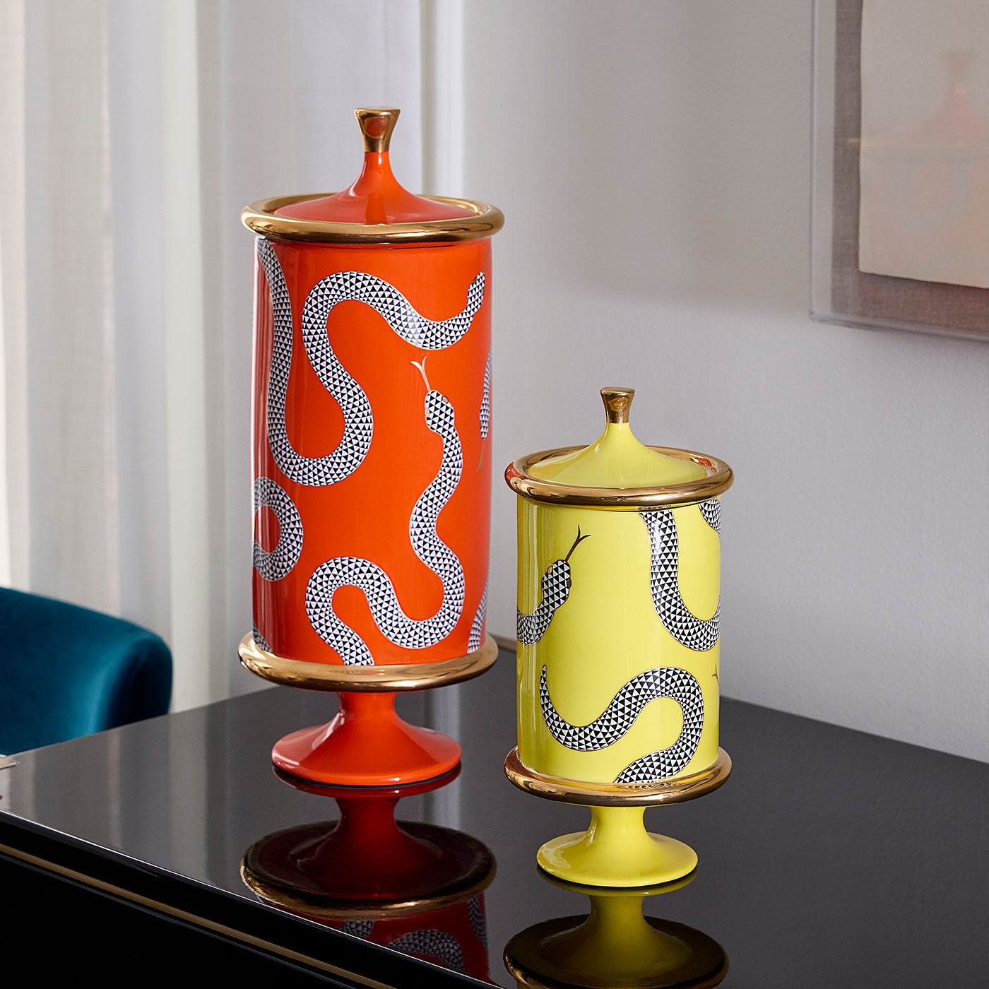 Two decorative ceramic lidded vases with snake motifs, one orange and one pale yellow, displayed on a glossy black table in an indoor setting.