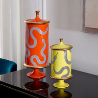 Two decorative ceramic lidded vases with snake motifs, one orange and one pale yellow, displayed on a glossy black table in an indoor setting.