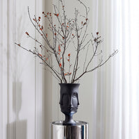A matte black ceramic vase shaped like a stylized female face, filled with dried branches and small orange berries, displayed on a reflective silver pedestal against a white paneled wall and curtain background.