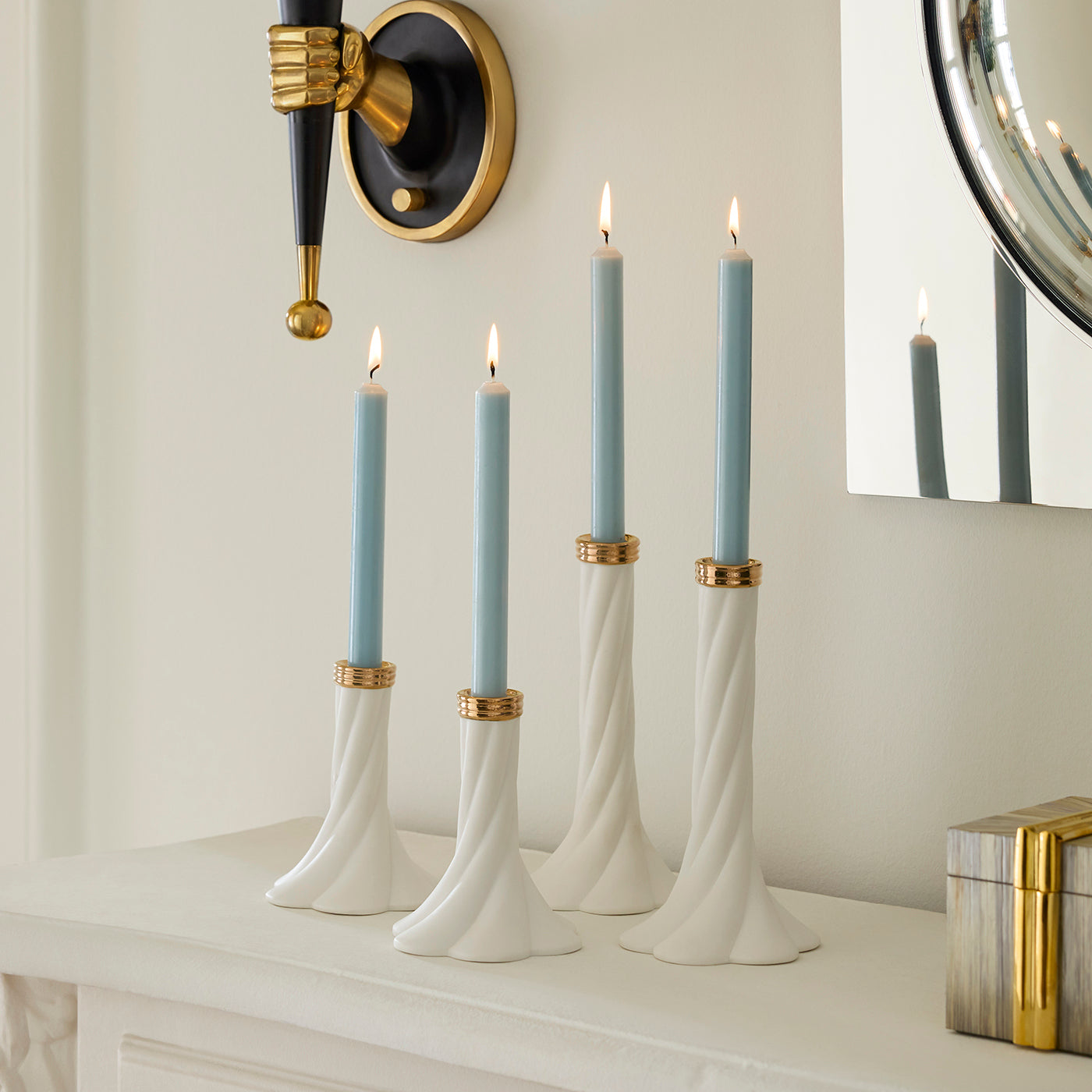 Four white twisted candlestick holders with gold accents holding light blue taper candles, styled on a white mantelpiece against a cream wall with a wall sconce and mirror visible.