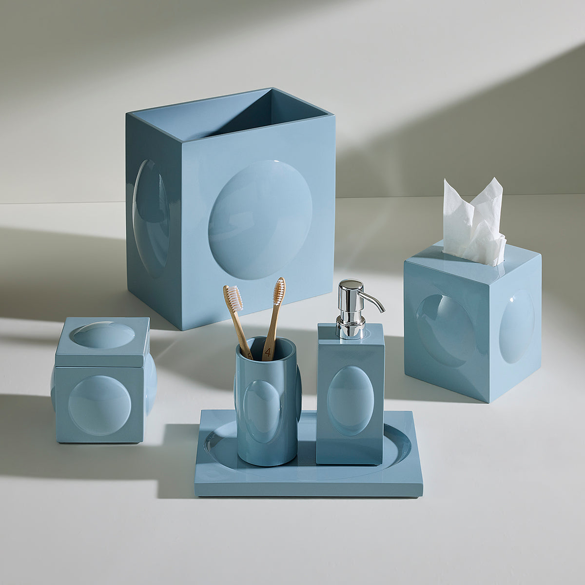 A coordinated bathroom accessory set in a matte dusty blue color, including a square tissue box cover with a tissue, a toothbrush holder, a soap dispenser, a cup, and a larger square organizer, arranged on a light neutral surface with soft shadows.