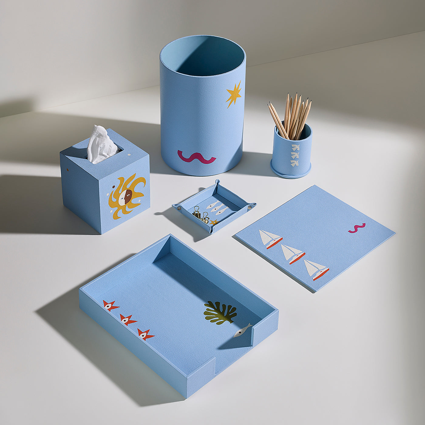 A coordinated light blue desk and bathroom accessory set arranged on a white surface, including a tissue box holder with a yellow sun, a cylindrical cup with a star, a small toothpick holder, a tissue box, a small tray, a rectangular tray, and a matching card or cover with sailboat motifs.