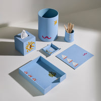 A coordinated light blue desk and bathroom accessory set arranged on a white surface, including a tissue box holder with a yellow sun, a cylindrical cup with a star, a small toothpick holder, a tissue box, a small tray, a rectangular tray, and a matching card or cover with sailboat motifs.