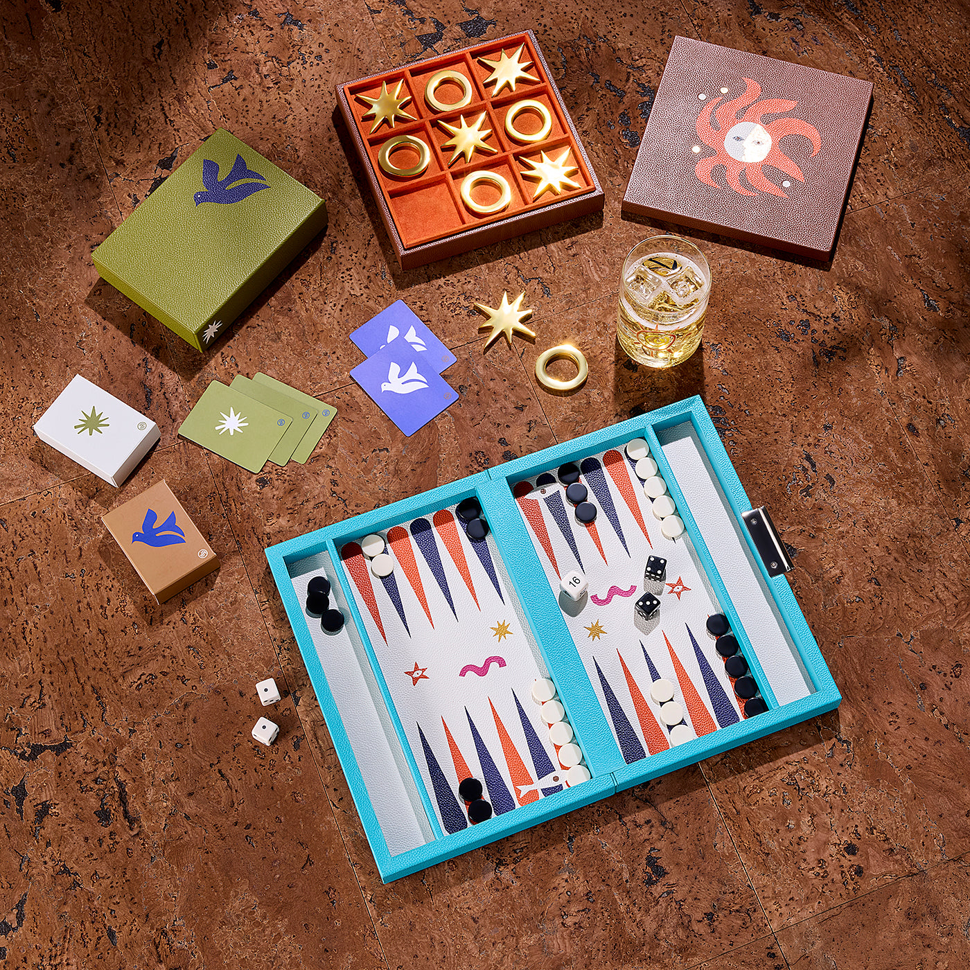 A styled flat-lay of a board game set on a textured brown surface, featuring a turquoise backgammon board, gold and white playing pieces, multiple colored dice, a glass of amber liquid, and several decorative boxes.