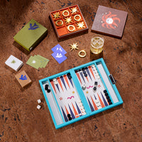 A styled flat-lay of a board game set on a textured brown surface, featuring a turquoise backgammon board, gold and white playing pieces, multiple colored dice, a glass of amber liquid, and several decorative boxes.