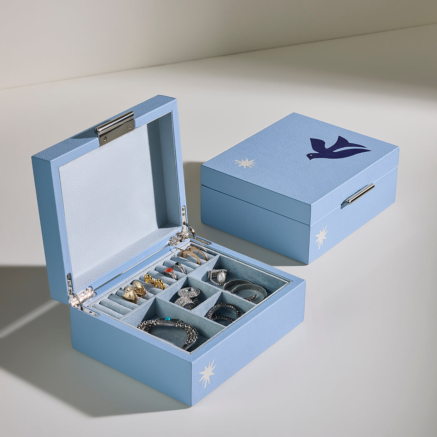 A light blue jewelry box shown open and closed on a neutral studio background. The open box contains multiple compartments holding rings and bracelets, and the closed box features a dark blue bird logo and small white starburst accents.