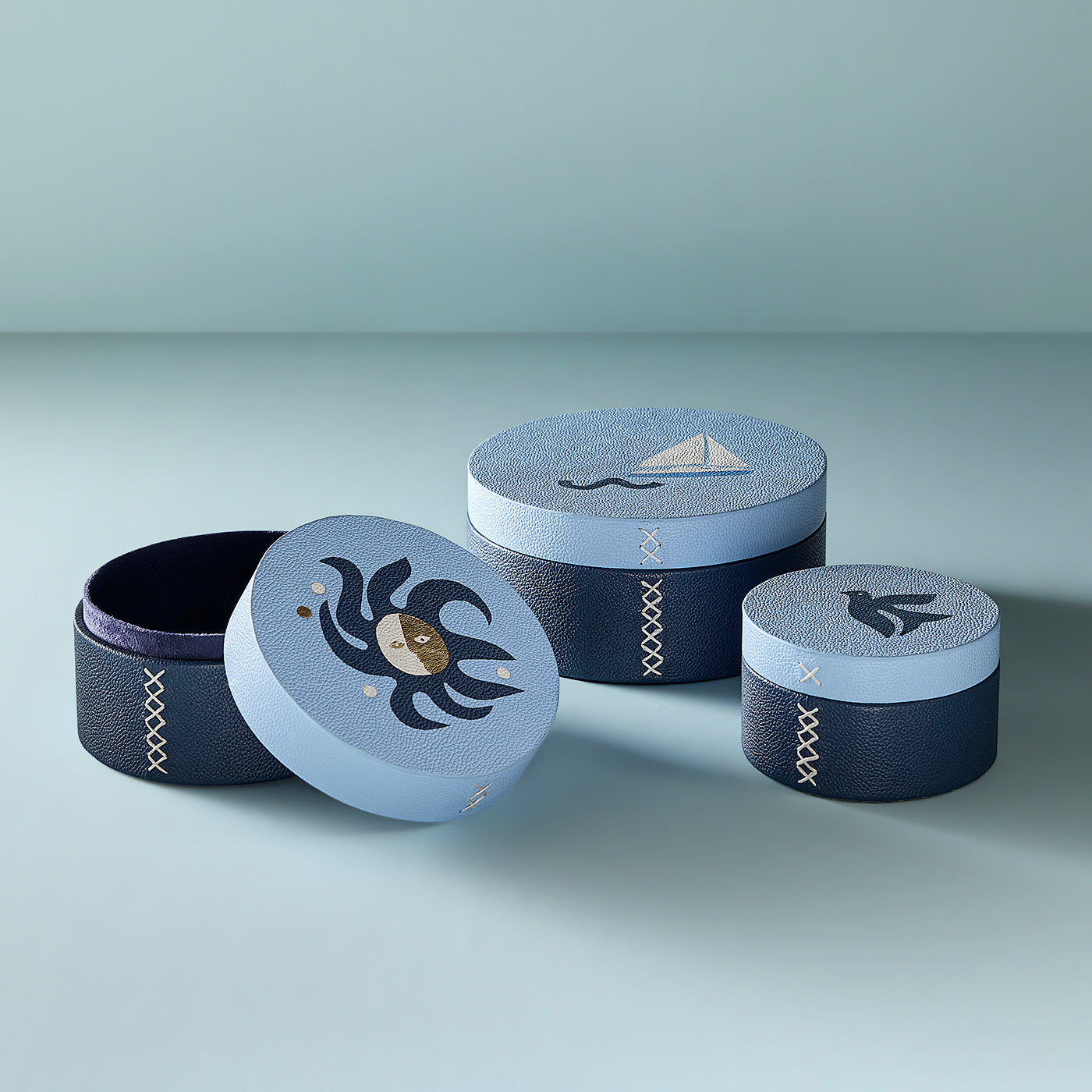 Three round decorative storage boxes with matching lids, arranged on a light gray surface against a pale blue-gray background. The boxes are dark navy with pale blue bands and embroidered details, including a crab motif on one lid, a sailboat on another, and a bird on the third.