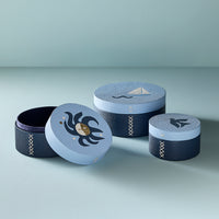Three round decorative storage boxes with matching lids, arranged on a light gray surface against a pale blue-gray background. The boxes are dark navy with pale blue bands and embroidered details, including a crab motif on one lid, a sailboat on another, and a bird on the third.