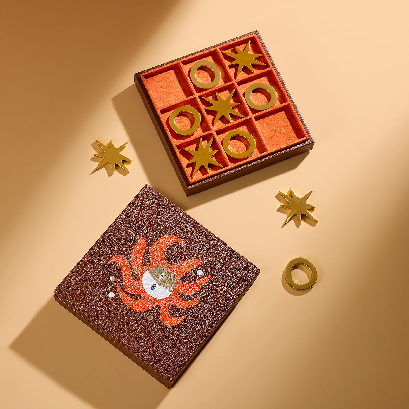 A styled flat-lay of a zodiac-themed game set on a warm beige background. The set includes a brown textured box with an orange fire sign illustration, a matching orange game board with gold-colored metal game pieces, and three gold star-shaped pieces.