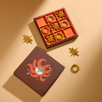 A styled flat-lay of a zodiac-themed game set on a warm beige background. The set includes a brown textured box with an orange fire sign illustration, a matching orange game board with gold-colored metal game pieces, and three gold star-shaped pieces.