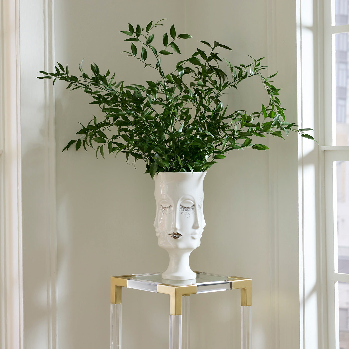 A white ceramic planter shaped like a stylized face with closed eyes, filled with leafy green branches, displayed on a glass and gold side table in a bright room.