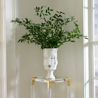 A white ceramic planter shaped like a stylized face with closed eyes, filled with leafy green branches, displayed on a glass and gold side table in a bright room.