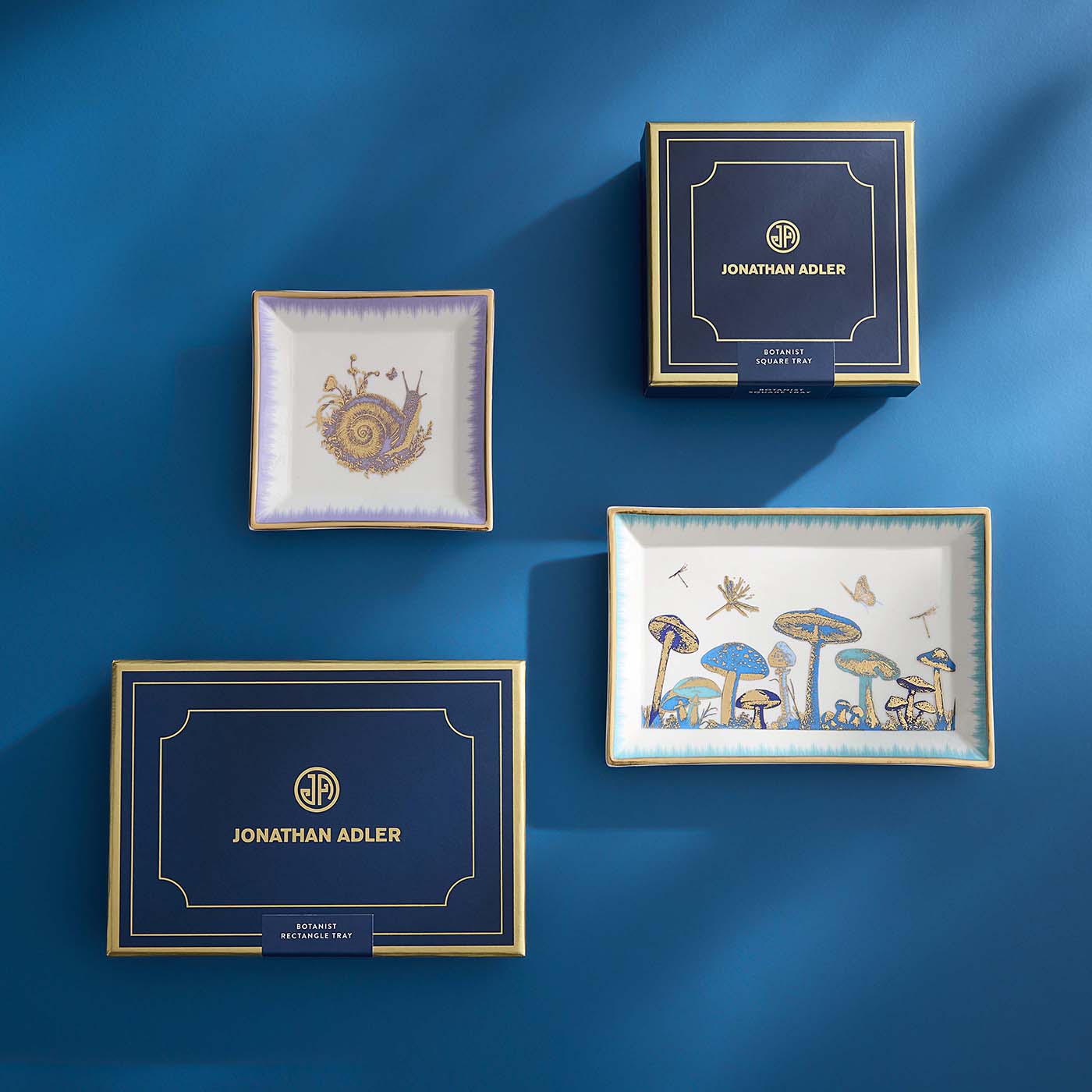 Decorative trays with artistic designs on a blue background, featuring the brand 'Jonathan Adler'.
